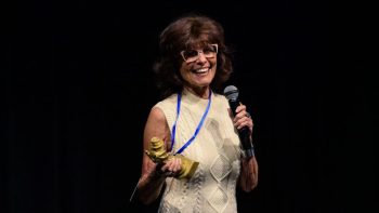 Adrienne Barbeau s'exprime devant le public du BIFFF ; elle porte de grandes lunettes et un pull blanc ; elle tient un micro dans la main gauche et une récompense dans la main droite (Chevalière de l'Ordre du Corbeau) ; photo prise au BIFFF 2026.
