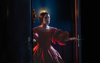 Une femme portant une belle robe de bal rouge ouvre une porte à double battant ; elle a les cheveux attachés et s'apprête à rentrer dans une pièce ; celle-ci est peu éclairée et une atmosphère victorienne se dégage de l'ensemble ; plan du film MARAMA de Taratoa Stappard.