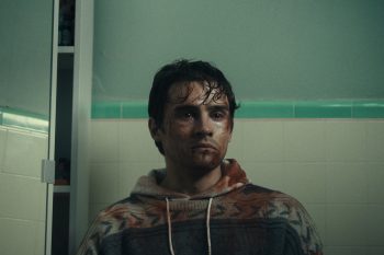 Un jeune homme aux cheveux noirs courts et au visage maculé de sang regarde fixement devant lui ; il porte un sweat en laine et à capuche, lui aussi taché de sang ; derrière lui, on devine les murs blancs et verts d'une salle de bain ; plan du film OBSESSION de Curry Barker.