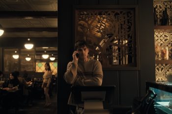 Un jeune homme est au téléphone dans un restaurant ; il se tient au milieu du plan, adossé à une sorte de panneau en bois ouvragé ; sur la gauche du plan, on aperçoit dans un léger flou la silhouette d'une jeune femme aux longs cheveux noirs, qui semble regarder dans la direction du panneau en bois ; au plafond, plusieurs lampes diffusent une lumière tamisée ; plan du film OBSESSION de Curry Barker.