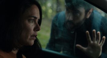 Une femme regarde dans le vide, enfermée dans l'habitacle de sa voiture ; un homme vient poser la main sur la vitre, en cherchant à capter son regard ; derrière lui, on aperçoit des bois ; plan du film SINGULAR, projeté au BIFFF 2026.