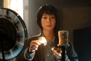 Une jeune femme asiatique semble mener un rituel ; elle tient une allumette enflammée dans la main droite et une sorte de talisman dans la main gauche ; elle s'approche d'une bougie située à l'avant-plan et reposant sur un pied ; sur la gauche de l'image, on devine la présence d'un cercle, qui est peut-être un cadran d'horloge ; la scène se passe de jour, à l'intérieur d'une pièce dont on ne perçoit que peu d'éléments ; plan du film NEVER AFTER DARK, projeté au BIFFF 2026.