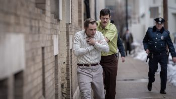 Un homme vêtu d'une chemise jaune et d'un pantalon marron contraint son otage à avancer dans la rue ; ce dernier porte une chemise blanche et un pantalon gris ; l'otage se tient le bas du cou, car on imagine qu'un fil en métal l'entrave ; derrière lui se tient donc son ravisseur, l'air déterminé ; en arrière-plan, on aperçoit un policier, qui suit la scène à distance ; on devine de la neige entassé sur le bas-côté de la rue dégagée ; plan du film LA CORDE AU COU de Gus Van Sant.
