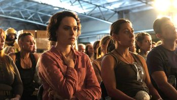 Des volontaires sont assis en rangs et écoutent ce qui doit être un briefing de mission ; la scène se passe dans un bâtiment de l'armée à en juger par les poutres métalliques au plafond ; Daisy Ridley est à l'avant-plan, le regard perdu dans le vide ; elle a la main droite posée à la base de son cou ; plan du film WE BURY THE DEAD, projeté au BIFFF 2026. 