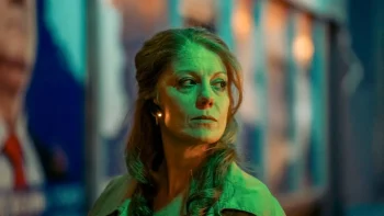 Une femme aux longs cheveux roux est filmée à hauteur d'épaule ; elle à la tête tournée vers la droite de l'image et regarde dans cette direction ; elle porte une oreillette à l'oreille droite, qui émet un signal lumineux ; derrière elle, on devine des fenêtre et des murs aux motifs bleus et orange ; plan du film KARMADONNA, projeté au BIFFF 2026.
