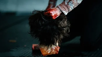 L'arrière d'une tête découpée est posée sur un sol sombre ; on n'aperçoit que les cheveux de la victime ainsi que du sang séché sur son cou ; une main ensanglantée saisit la tête par les cheveux ; la main est celle d'un homme qu'on devine accroupi dans la pièce ; sa chemise est tachée de sang séché ; plan du film INCOMPLETE CHAIRS, projeté au BIFFF 2026.