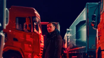 De nuit, un homme en manteau noir se tient devant des camions ; on aperçoit l'avant d'un camion rouge vif sur la gauche et l'arrière d'un autre camion, bleu celui-là, sur la droite ; plan du film DU FIOUL DANS LES ARTERES, présenté à Cannes 2026.