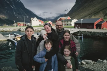 Une famille pose devant un fjord norvégien ; le père a le crâne chauve et porte des lunettes, la mère sourit et tient leur bébé emmitouflé dans ses bras ; quatre autres enfants se tiennent autour de leurs parents ; en arrière-plan, on aperçoit des bâtiments sur les berges d'un fjord, avec des montagnes situées au loin ; plan du film FJORD, présenté à Cannes 2026.