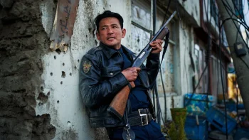 hope-na-hong-jin-policier-cannes-2026 Un homme asiatique est en uniforme et policier et tient une carabine entre les mains ; il serre les dents en regardant vers la gauche du cadre, tout en ayant le dos appuyé contre le mur blanc d'une maison ; plan du film HOPE, présenté à Cannes 2026.