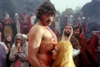 Un homme moustachu et torse nu porte une couronne d'épines ; il a le torse couvert du sang qui s'écoule depuis son crâne et regarde vers le bas ; à ses pieds se tient une femme blonde, la tête posée contre le ventre de l'homme, qui pourrait être Jésus ; derrière eux, une assemblée d'individus est tournée dans leur direction ; plan du film LES DIABLES, présenté à Cannes 2026.