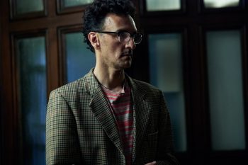 Le réalisateur Taratoa Stappard se tient debout et porte une veste à petits carreaux ; il porte des lunettes et regarde vers la droite de l'image ; derrière lui, on aperçoit des montants en bois ; plan du film TAUMANU.