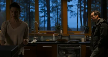 Un homme et une femme se tiennent dans une cuisine sombre ; la femme est affairée avec de la vaisselle sur la gauche du plan ; l'homme se tient adossé sur la droite du plan, le regard dans le vide ; tous deux semblent distants l'un de l'autre ; derrière la baie vitrée de la cuisine, on aperçoit des troncs d'arbres et un ciel entre chiens et loups ; plan du film MINOTAURE, présenté à Cannes 2026.