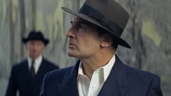 moulin-gilles-lellouche-nemes-cannes-2026 Un homme portant un chapeau gris regarde sur sa droite, en levant légèrement la tête ; l'homme est joué par Gilles Lellouche ; il est vêtu d'une chemise blanche et d'un veston bleu marine ; derrière lui apparaît la silhouette floue d'un autre homme à chapeau et en costume ; plan du film MOULIN, présenté à Cannes 2026.