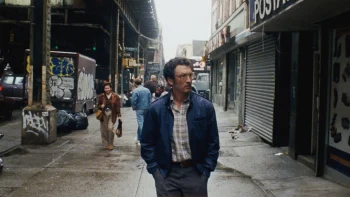 paper-tiger-james-gray-rue-cannes-2026 Un homme à lunettes, vêtu d'une chemise à carreaux et d'une veste bleue, marche dans une rue ; il est à l'avant-plan et, derrière lui, on aperçoit du mobilier urbain couvert de graffitis, de même que des passants ; les devantures des magasins sont fermées, avec les volets clos ; plan du film PAPER TIGER, présenté à Cannnes 2026.