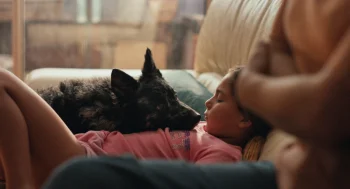 Un enfant en t-shirt rose est affalé dans un divan blanc en cuir ; un chien noir repose allongé sur sa poitrine ; à l'avant-plan, on devine la présence d'une autre personne, les bras croisés ; plan du film QUELQUES MOTS D'AMOUR, présenté à Cannes 2026.