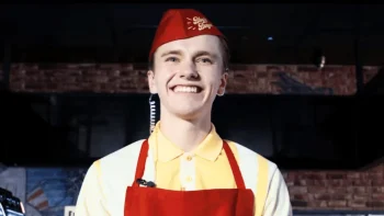 Un employé de fast-food sourit à pleines dents ; il porte un képi rouge avec l'enseigne de son entreprise écrite en jaune ; le fil d'une oreillette pend de son oreille gauche ; il porte une chemise jaune et un tablier rouge ; plan du film SANGUINE, présenté à Cannes 2026.