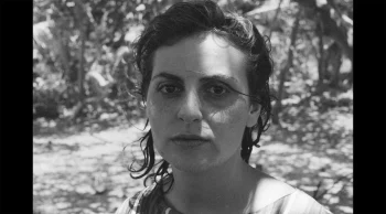Le visage d'une femme en noir et blanc, au regard perdu dans le vide en direction de la caméra ; il s'agit du visage de Michèle Firk ; derrière elle, on devine un jardin, avec des arbres ou de la végétation haute ; plan du film UNE VIE MANIFESTE, présenté à Cannes 2026.