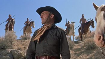 Plan en contre-plongée sur Randolph Scott, debout, qui ne voit pas quatre Amérindiens derrière lui, sur leurs chevaux dans le film Comanche Station de Buedd Boetticher, figurant dans Trois maîtres du cinéma modeste.