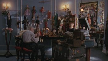 André Toulon, vu de dos, assis à sa table de travail, retouche une poupée ; sur les murs, tout autour, de très nombreuses autres poupées sont accrochées ; plan issu de Puppet Master (1989).