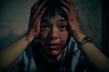 Une jeune femme eurasienne se tient la tête entre les mains ; elle a des cheveux noirs et des yeux noirs ; elle porte une montre au poignet droit ainsi qu'un t-shirt bleu ; ses mains et une partie de son visage sont recouverts de traces de sang, tandis qu'elle affiche un air désemparé ; derrière elle, on devine un mur gris, mais la pièce où le personnage se trouve est plongée dans une luminosité faible ; plan du film SACCHARINE de Natalie Erika James.