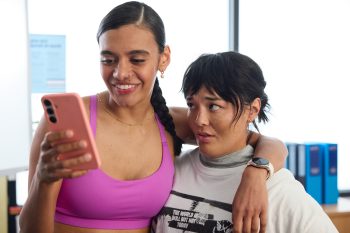 Une jeune femme métisse regarde l'écran de son téléphone en souriant, tandis qu'elle a le bras droit autour du cou d'une autre jeune femme, eurasienne ; la jeune femme qui regarde son téléphone porte un top rose et a les cheveux coiffés en une tresse ; l'autre personnage féminin porte un t-shirt blanc et affiche une mine inquiète ; derrière les deux jeunes femmes, on aperçoit des murs blancs, ainsi que des classeurs rangés sur une armoire basse ; plan du film SACCHARINE de Natalie Erika James. 