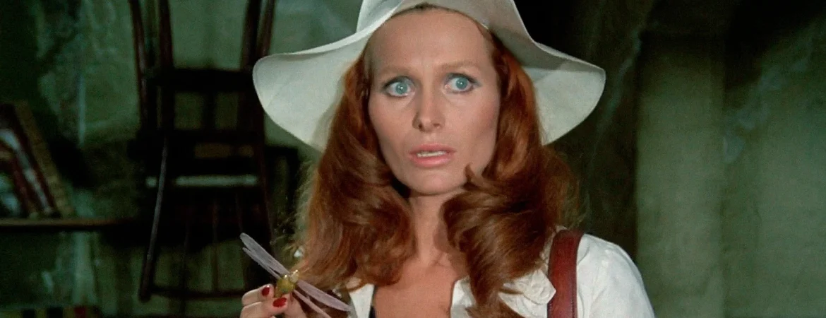 Une femme rousse habillée en blanc et avec un chapeau blanc tenant dans sa main une fausse libellule dans un décor verdâtre avec des chaises en arrière plan.