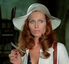 Une femme rousse habillée en blanc et avec un chapeau blanc tenant dans sa main une fausse libellule dans un décor verdâtre avec des chaises en arrière plan.