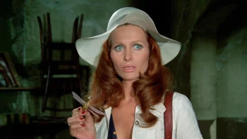 Une femme rousse habillée en blanc et avec un chapeau blanc tenant dans sa main une fausse libellule dans un décor verdâtre avec des chaises en arrière plan.