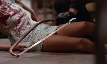 Homme ganté tenant une scie à métaux au dessus du corps d'une jeune femme en robe blanche, couverte de sang.