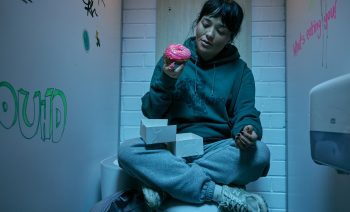 Une jeune femme eurasienne sort un donut au nappage rose d'une boîte en carton, qu'elle regarde avec envie ; elle est assise en tailleur sur des toilettes, lunette baissée ; les jambes croisées, elle porte un sweat à capuche vert foncé, un pantalon de training gris et une paire de baskets blanches ; les murs des toilettes sont blancs et quelques tags recouvrent les cloisons latérales ; plan du film SACCHARINE de Natalie Erika James.