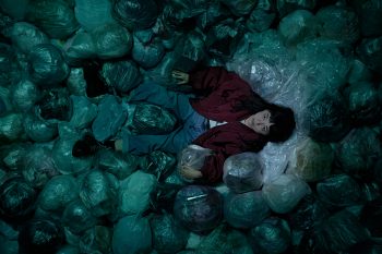 Une jeune femme filmée en plan zénithal est allongée au milieu de sacs poubelle de couleur verte ; elle a des cheveux noirs courts et porte un gilet foncé sur un vêtement blanc ; elle regarde vers le haut, tout en étant cernée de sacs ; plan du film SACCHARINE de Natalie Erika James.