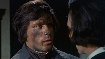 Deux hommes se font face et se regardent, l'un tournant le dos à la caméra, l'autre ayant le visage couvert de suie ; ce dernier a les cheveux mi-longs et la lèvre inférieure ensanglantée ; l'homme de dos a des cheveux noirs, un col blanc et probablement un vêtement noir ; plan du film LE FASCINANT CAPITAINE CLEGG de la Hammer.