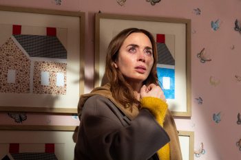 Une femme, jouée par Emily Blunt, est dans ce qui s'apparente à une chambre d'enfants ; elle regarde vers le haut et a le visage plongé dans une lumière qui vient probablement du plafond, ou de l'extérieur ; elle porte un pull jaune et un manteau vert foncé ; derrière elle, on aperçoit quatre cadres suspendus au mur représentant des maisons ; le papier peint évoque celui d'une chambre d'enfants ; plan du film DISCLOSURE DAY de Steven Spielberg.