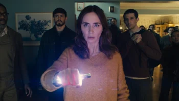 Une femme, jouée par Emily Blunt, serre un étrange artéfact lumineux dans une main, qu'elle tend en direction de la caméra ; elle porte de longs cheveux foncés et est vêtue d'un pull rouge ou rose ; derrière elle, on aperçoit plusieurs hommes, qui regardent tous dans la même direction qu'elle (vers la caméra) ; tous ont l'air subjugués par ce qu'ils voient ; la scène se déroule dans la pièce d'une maison ; sur un des murs, on aperçoit un tableau ; plan du film DISCLOSURE DAY de Steven Spielberg.