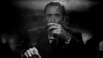 Un homme, joué par Nicolas Cage, porte un verre de vin à sa bouche ; dans l'autre main, il tient une cigarette fumante ; l'arrière-plan est trop sombre pour que l'on puisse distinguer quoi que ce soit ; de la fumée s'est agglutinée au-dessus de la tête du personnage, en costume noir ; l'image est en noir et blanc ; plan de la série SPIDER-NOIR de Prime Vidéo. 