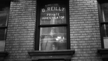 Façade d'un bâtiment en brique, percé par une fenêtre en son centre ; sur le haut de la fenêtre, il est écrit "B. Reilly, Private Investigator" ; de l'autre côté de la fenêtre, on devine une silhouette d'homme en costume ; deux autres fenêtres encadrent la première, sur sa gauche et sur sa droite, mais elles ne sont que très partiellement visibles ; l'image est en noir et blanc ; plan de la série SPIDER-NOIR de Prime Vidéo.