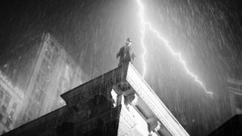 Spider-Noir, filmé en contre-plongée, se tient debout au bord du toit plat d'un building ; on aperçoit d'autres hauts immeubles en arrière-plan, tandis qu'il pleut abondamment sur la ville ; un éclair lumineux zèbre le ciel noir ; l'image est en noir et blanc ; plan de la série SPIDER-NOIR de Prime Vidéo.