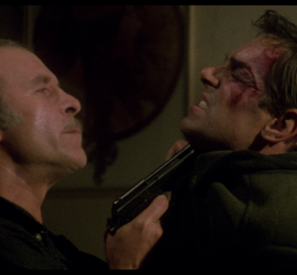Un gangster tient Fabio Testi par le col, le menaçant avec son revolver, dans le film La guerre des gangs de Lucio Fulci.