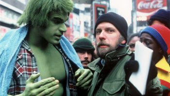 Lou Ferrigno sur le tournage de la série "L'Incroyable Hulk" ; il est à gauche du plan, en géant vert, portant une chemise bariolée ouverte et une serviette de bain sur les épaules ; à ses côtés se tient un homme barbu et moustachu, qui doit être le réalisateur ; l'homme porte un bonnet noir et tend sa main ganté vers la caméra, tout en tenant un carnet ou une feuille de papier pliée ; derrière eux, on aperçoit plusieurs personnes, sans doute des acteurs ou des figurants, de même que des rayons d'un supermarché ; plan de la série L'INCROYABLE HULK, rééditée par Elephant Films.