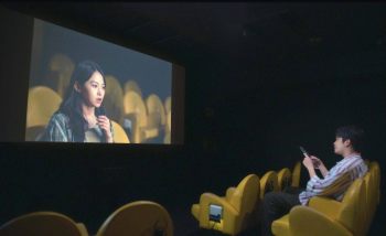 Un homme assis dans une salle de cinéma aux fauteuils jaunes, regarde sur l'écran une femme elle même assis dans une salle de cinéma à fauteuils jaunes