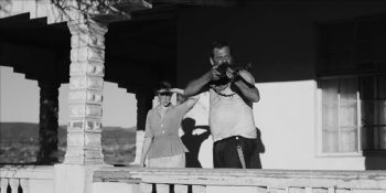 Un homme met en joue quelque chose ou quelqu'un hors champs devant le regard d'une femme derrière lui