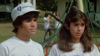 Deux adolescents sont côte à côte dans ce qui s'apparente à un jardin ; à gauche du plan, le garçon aux cheveux bruns porte un bob blanc et un t-shirt blanc ; à droite du plan, la jeune fille aux longs cheveux châtains porte également un t-shirt blanc ; en arrière-plan, on devine d'autres enfants en t-shirt blanc peut-être en train de monter une grande tente ; plan du film MASSACRE AU CAMP D'ETE, édité par ESC.