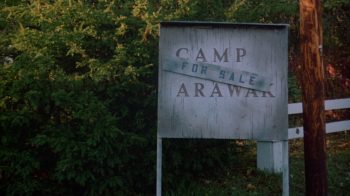 Un panneau en bois indiquant l'entrée du "Camp Arawak" sur lequel est collé un plus petit écriteau "for sale" ; le panneau est blanc, mais usé par le temps ; derrière lui, on aperçoit des buissons ou des arbustes, ainsi qu'une barrière blanche ; plan du film MASSACRE AU CAMP D4ETE, édité par ESC. 