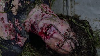 La tête et le buste ensanglantés d'une personne couchée au sol, de la bouche de laquelle s'échappe un serpent ou un reptile ; les yeux du mort sont figés et du sang tache son visage et le haut de son corps sans vie ; plan du film MASSACRE AU CAMP D'ETE, édité par ESC.