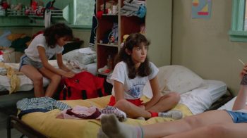 Une adolescente aux longs cheveux bruns est assise en tailleur sur un lit ; elle porte un short rouge et un t-shirt blanc et semble regarder dans le vide ; le drap de son lit et jaune ; on comprend qu'il doit s'agir d'un dortoir ; derrière elle, une autre jeune fille est assise sur un lit et paraît fouiller dans son propre sac de couleur rouge ; en amorce du plan, on aperçoit deux jambes étendues, qui indiquent la présence d'une troisième personne et d'un troisième lit ; plan du film MASSACRE AU CAMP D'ETE, édité par ESC.