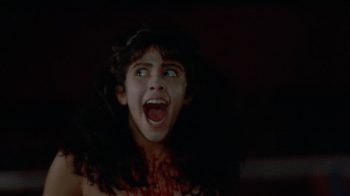 Dans la nuit noire, une adolescente semble hurler, la bouche grande ouverte et les yeux tournés vers la gauche du plan ; elle porte de longs cheveux foncés qui recouvrent ses épaules ; plan du film MASSACRE AU CAMP D'ETE, édité par ESC.