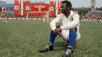 Un joueur en maillot blanc est assis sur la pelouse d'un stade de foot ; entouré de banderoles avec des crois gammées.