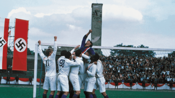 escape-to-victory-john-huston Sylvester Stallone soulevé par son équipe près d'un but de football ; ses équipiers portent des maillots et des shorts blancs ; sur la gauche, en arrière-plan, planent des drapeaux nazis ; on aperçoit également les gradins remplis de supporteurs ; plan du film ESCAPE TO VICTORY de John Huston.