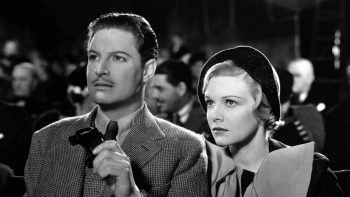 Un homme élégant, joué par Robert Donat, est aux côtés d'une belle femme blonde, jouée par Madeleine Carroll, au sein du public d'un spectacle ; ils sont filmés en plan rapprochés et ont le regard inquiet, les yeux rivés devant eux, sans doute vers la scène ; l'homme a les cheveux gominés et une petite moustache ; il tient une petite paire de jumelles dans une main ; la femme a l'air grave et concentrée, portant un béret noir ; derrière eux, on aperçoit le reste de l'audience, essentiellement des hommes assis ; plan du film LES 39 MARCHES, réédité par Elephant Films.