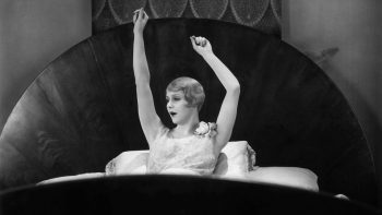 Une jeune femme blonde semble se réveiller dans un grand lit ; elle s'étire en levant les bras, le dos appuyé contre son oreiller ; la tête de lit est proéminente, il s'agit d'un grand demi-cercle sombre et classieux ; plan du film LE PASSE NE MEURT PAS, réédité par Elephant Films.